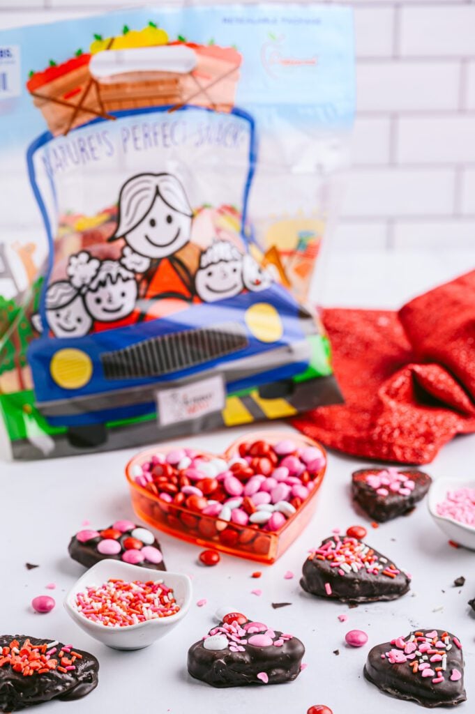 vertical shot of chocolate covered hearts with m&ms and sprinkles and bag of The Produce Moms apples in background