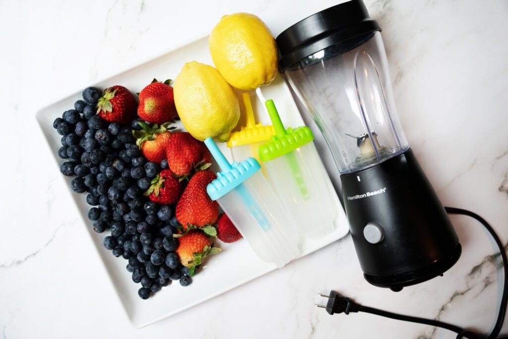 Ingredients for Bomb Pop Fruit Popsicles (strawberries, blueberries and lemons on a plate with blender)