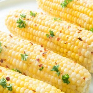 close up vertical shot of boiled corn on the cob on white plate with red pepper flakes and parsley