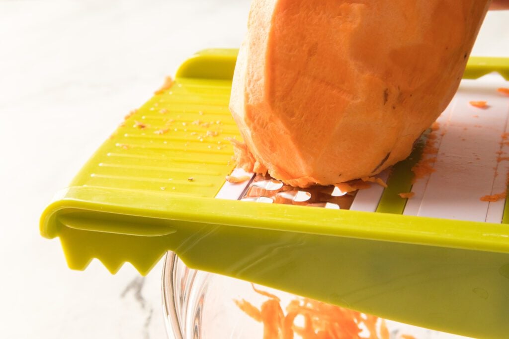 shredding sweet potato on a grater over a glass bowl