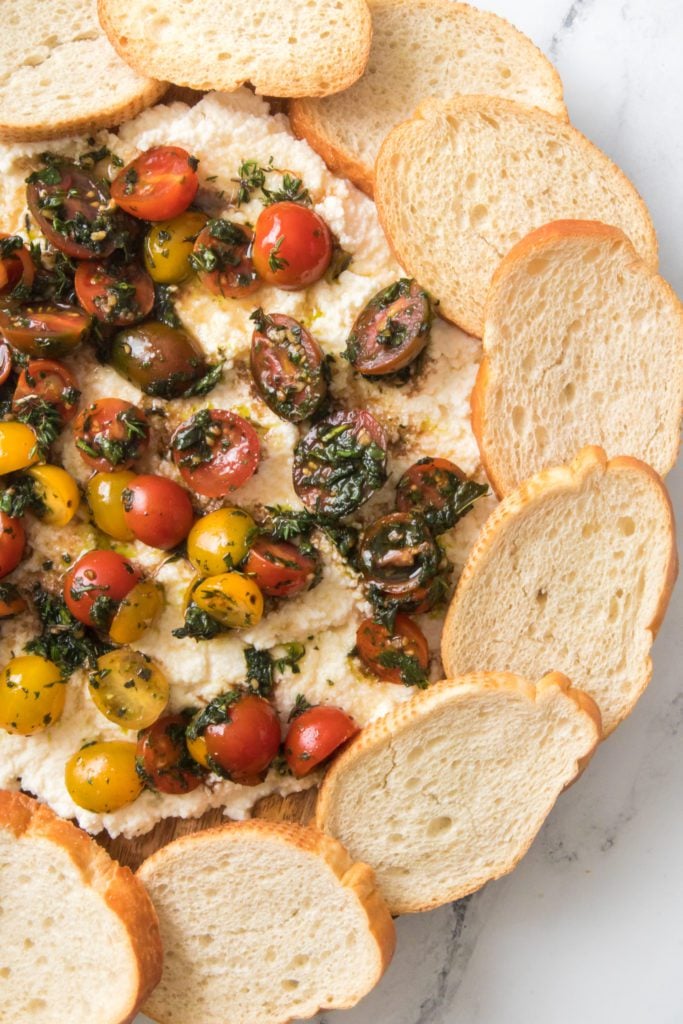 close up vertical shot of round charcuterie board with ricotta, fresh tomatoes and herbs along with bread for serving