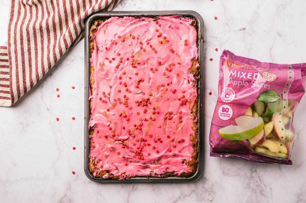 overhead shot of apple slices on baking sheet with pink glaze and heart sprinkles with a bag of crunch pak apple slices