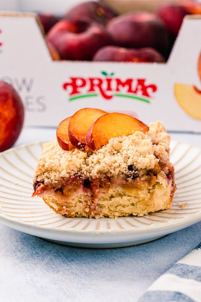 peach coffee cake on plate topped with fresh peaches and box of prima peaches in the background