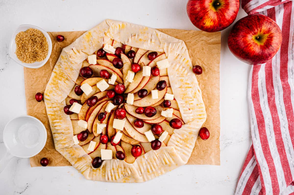 overhead shot of apples placed in a circle pattern in pie crust, topped with butter slices and fresh cranberries
