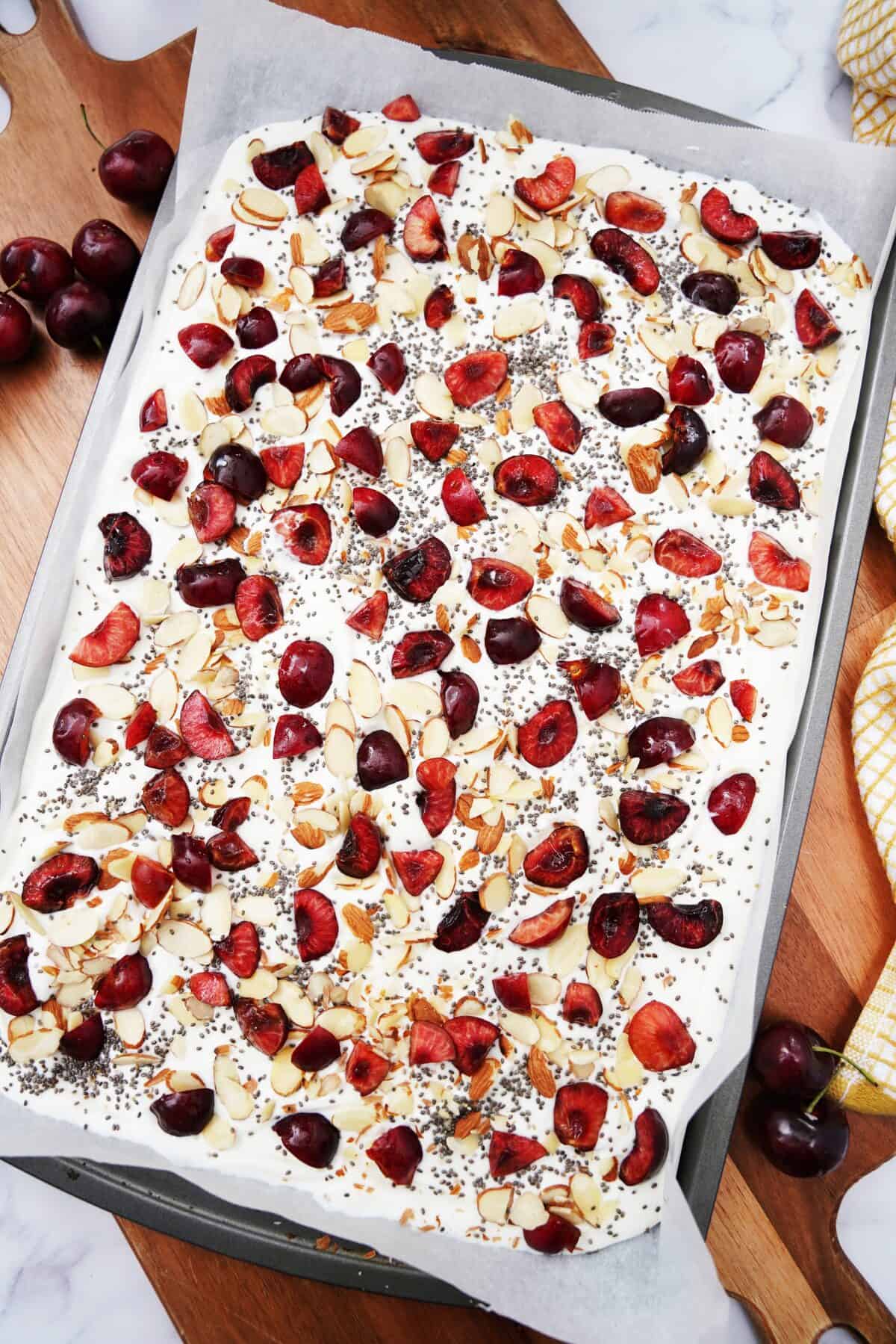 overhead shot of greek yogurt spread into an even layer on a baking sheet topped with sliced almonds, chia seeds and cherries