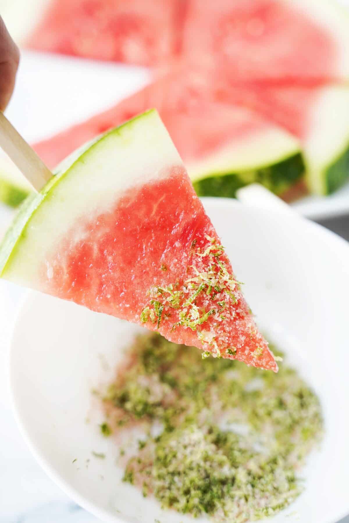 hand holding popsicle stick with a slice of watermelon dipped in tequila topped with salt and lime zest