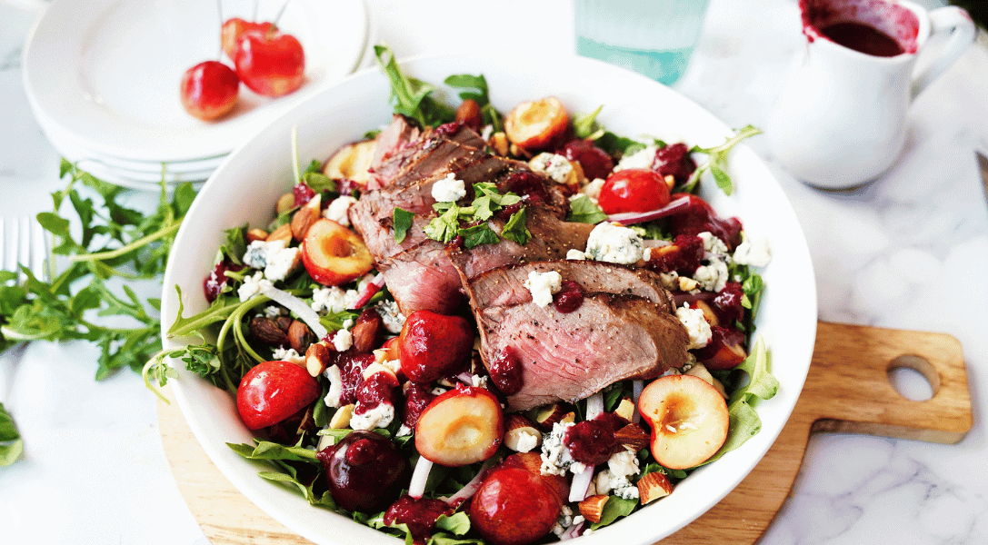 Steak & Arugula Salad with Cherries and Almonds in a white bowl surrounded by cherries