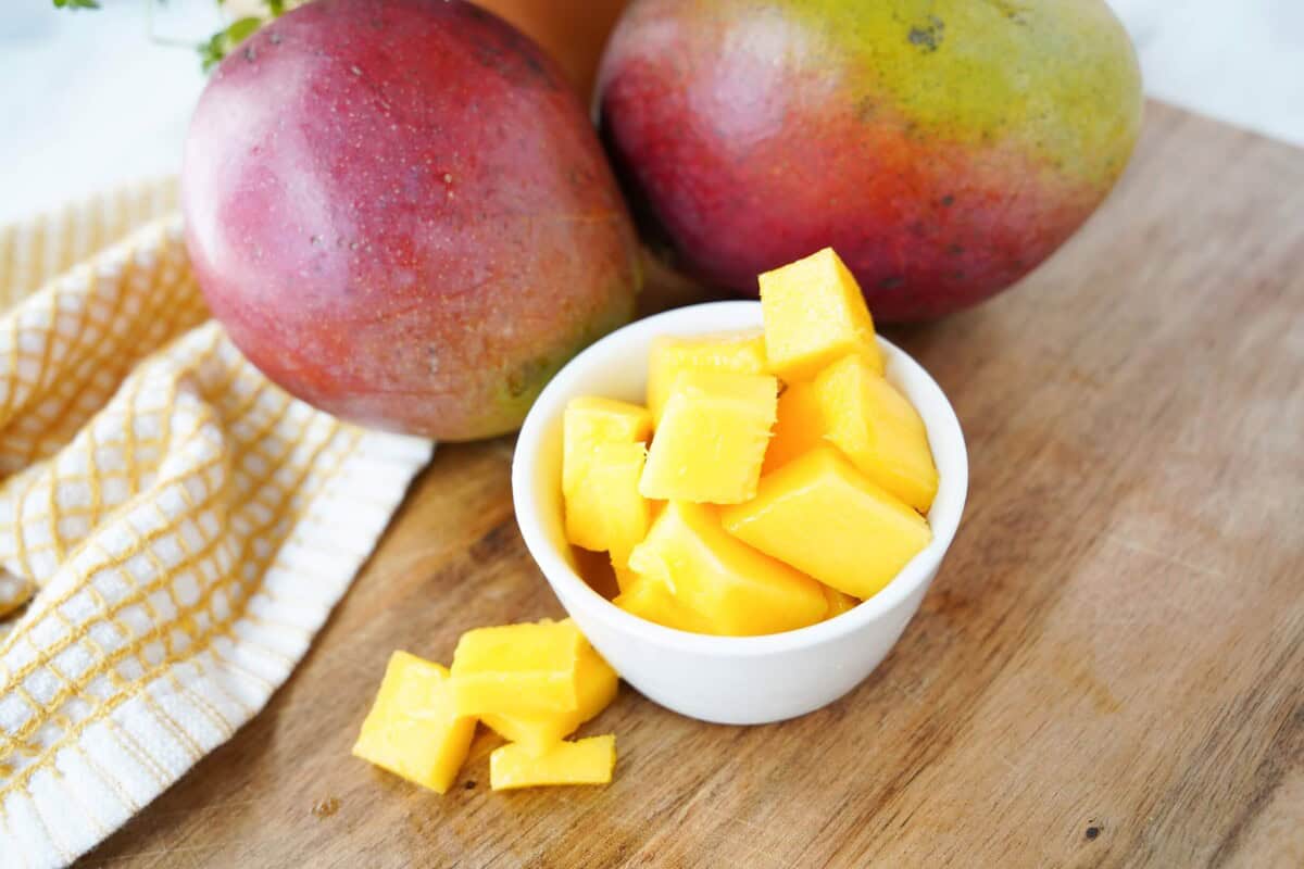 Cubed mango in a white bowl with 2 mangoes behind it