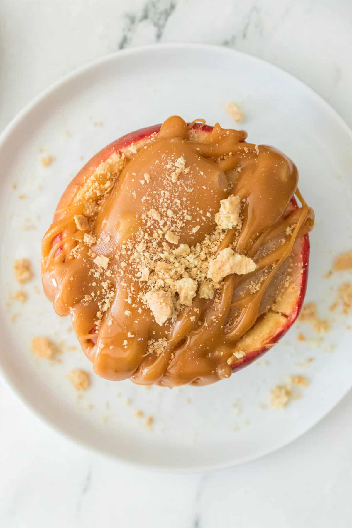overhead shot of stuffed baked apple with cheesecake filling on white plate topped with caramel sauce and crushed graham crackers