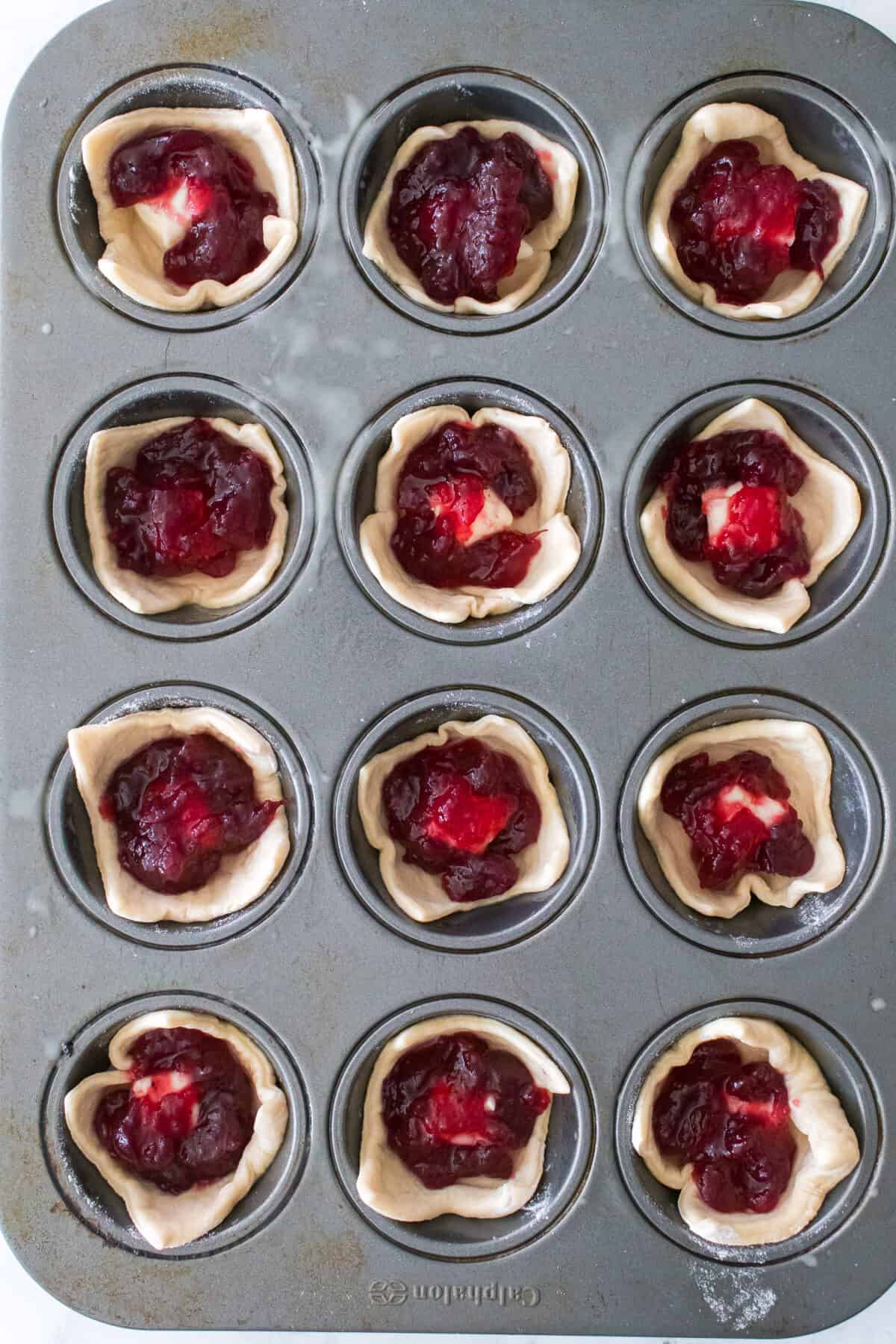 muffin tin with crescent dough pieces, brie cheese and cranberry sauce prior to baking