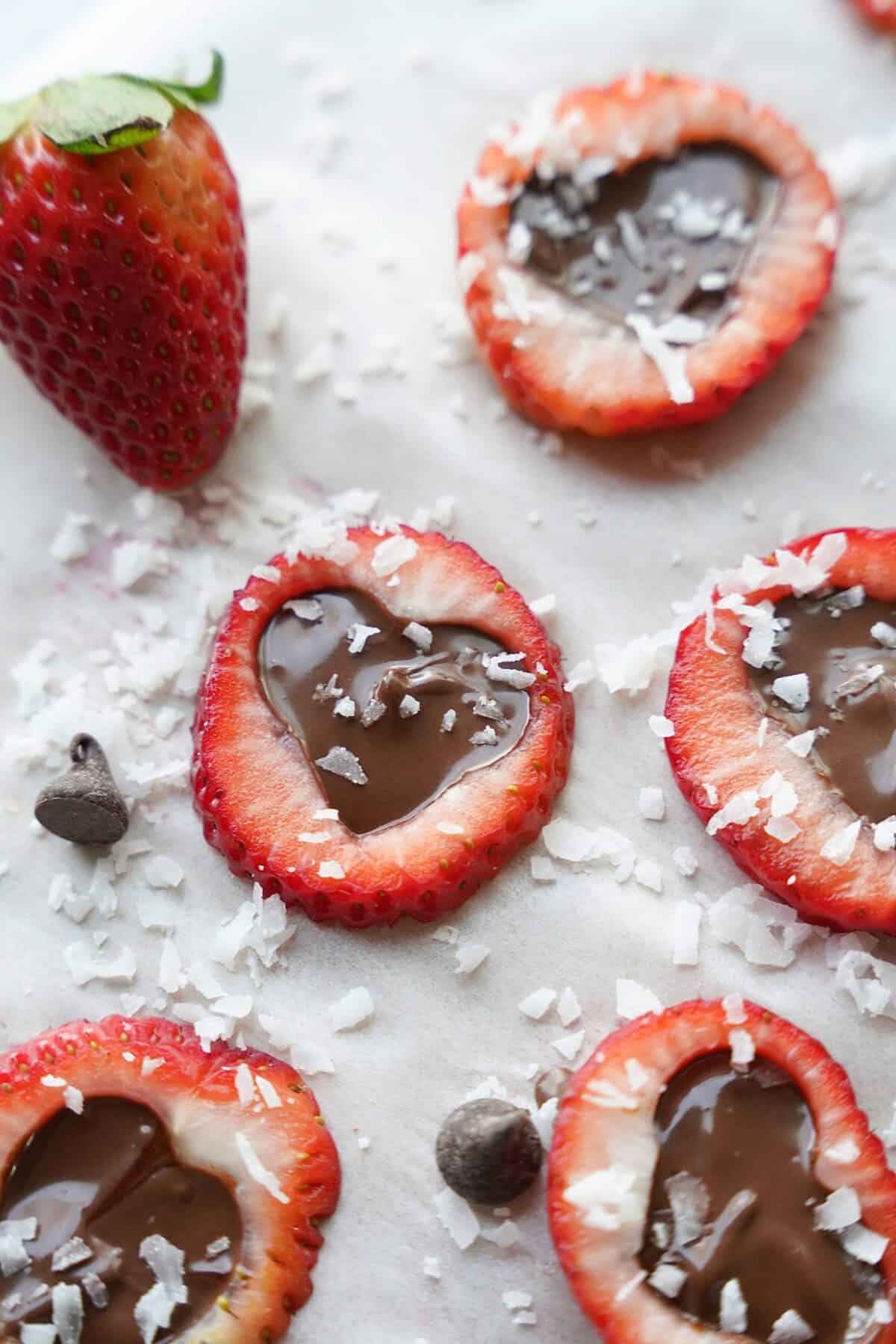 overhead shot of sliced strawberries on parchment paper with heart cut out, chocolate in the center shaped as a heart and topped with chopped coconut flakes