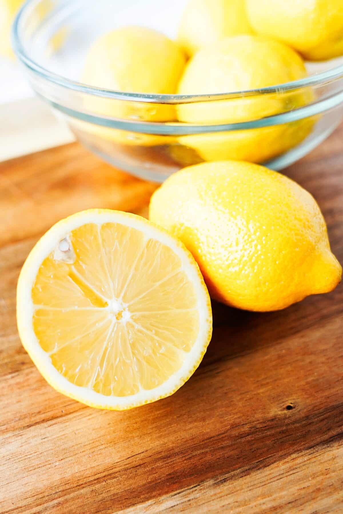 wood cutting board with bowl of lemons in background and lemon in front cut in half