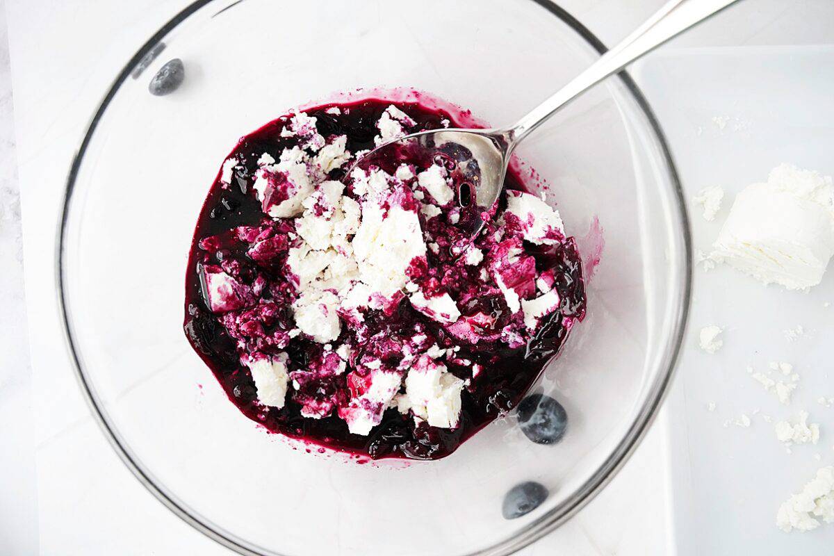 goat cheese being added to blueberry sauce in glass bowl with spoon