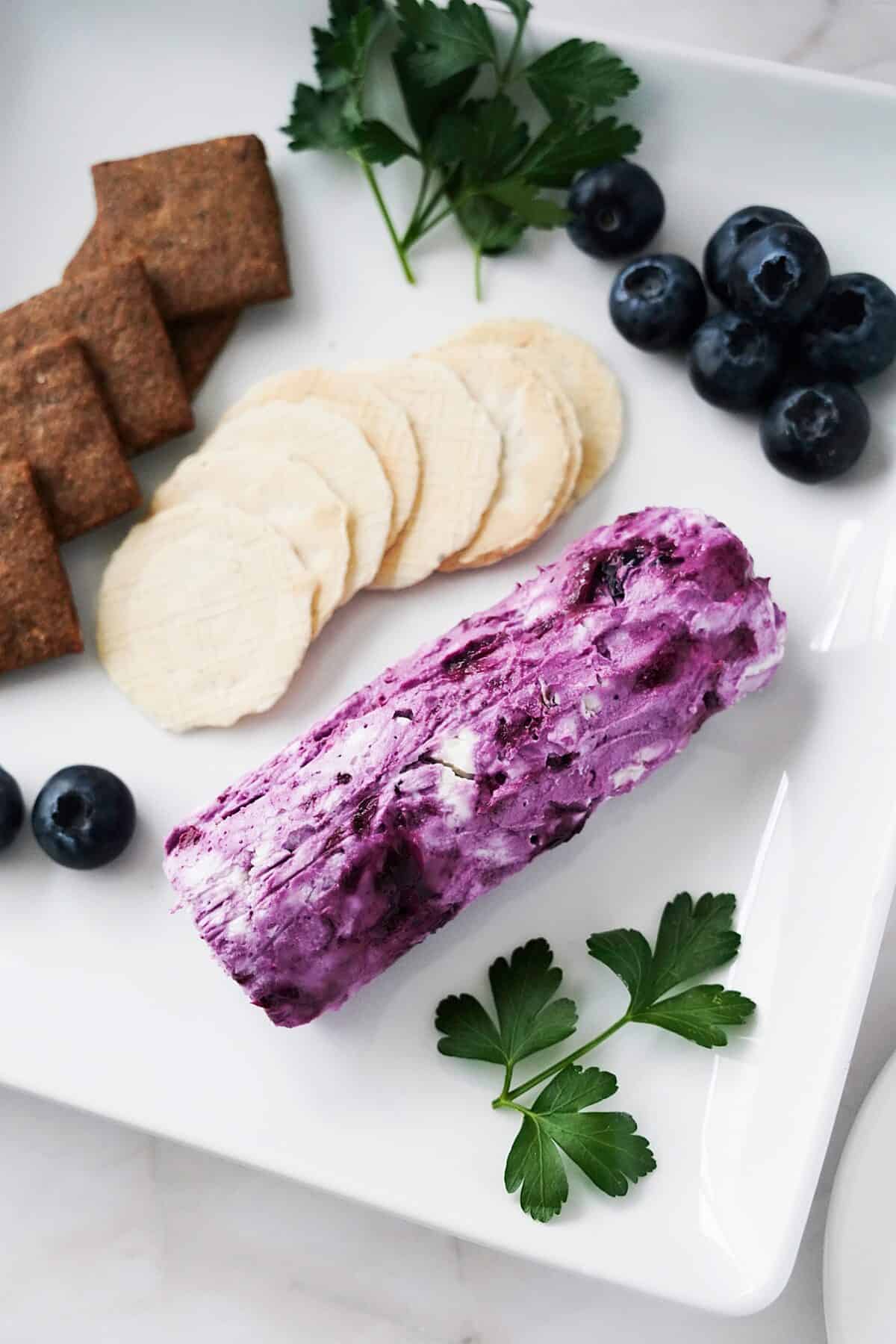 vertical shot of blueberry goat cheese log on serving platter with crackers, blueberries and stems of parsley