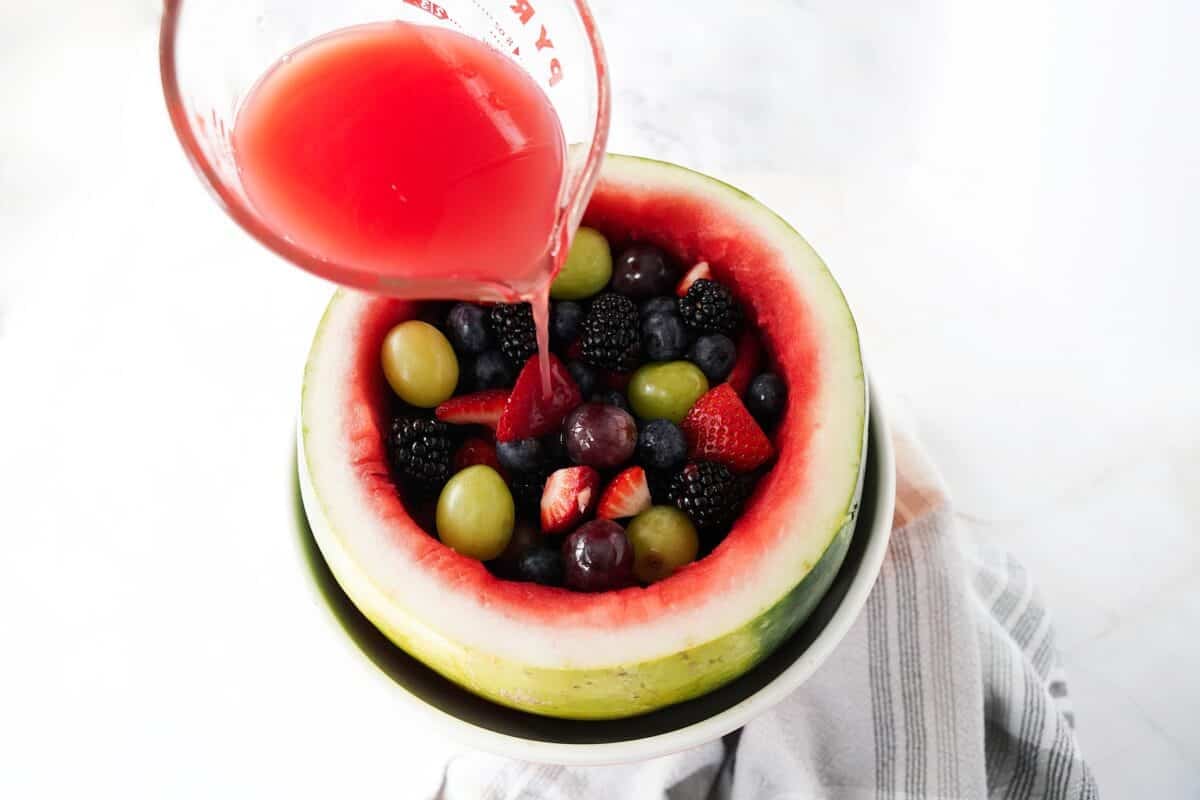 gelatin being poured from measuring cup into watermelon with fresh fruit