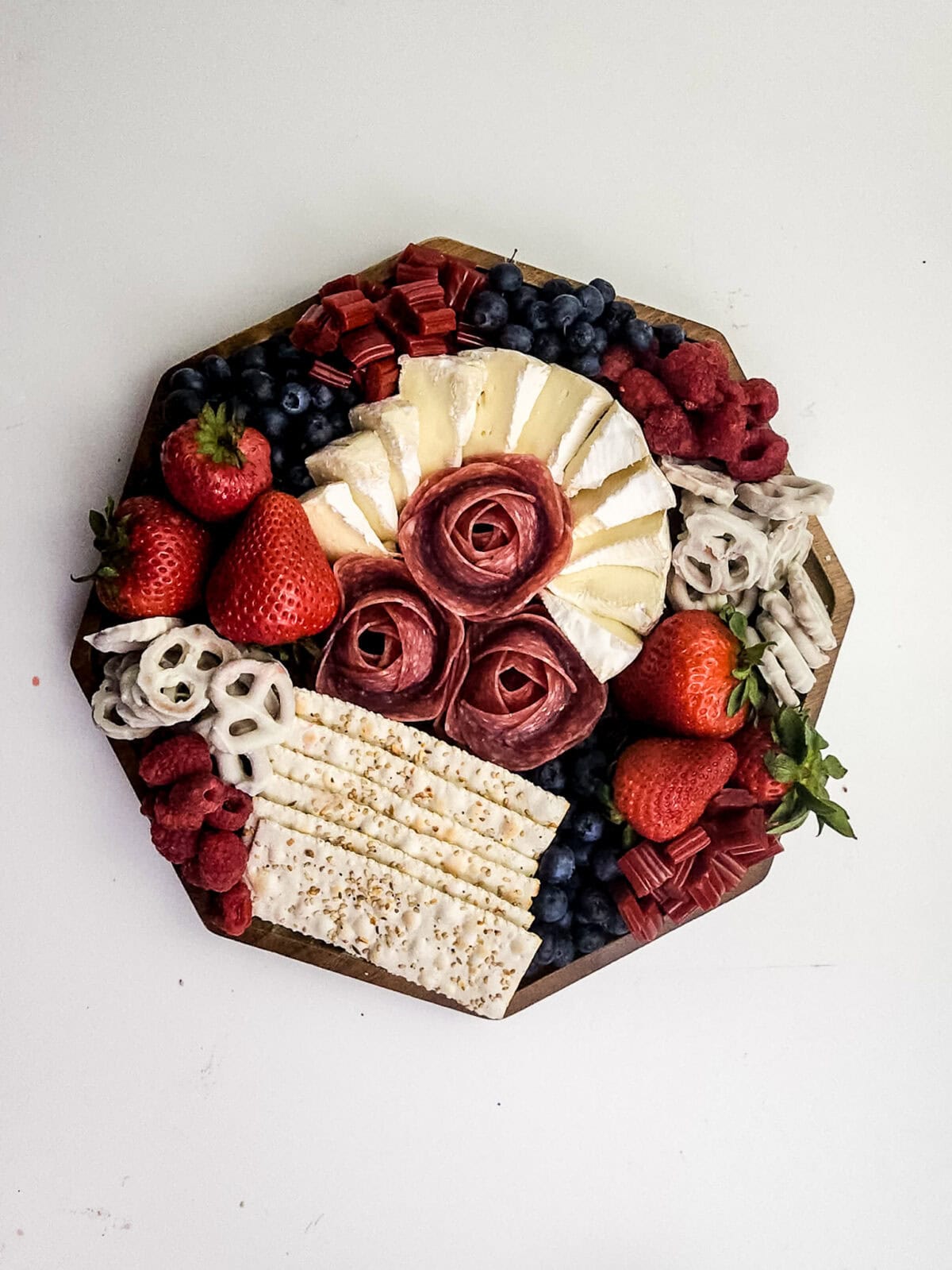 overhead shot of crackers, salami rosettes, brie cheese, blueberries,  strawberries, raspberries, white chocolate pretzels, twizzler pieces and raspberries on cutting board