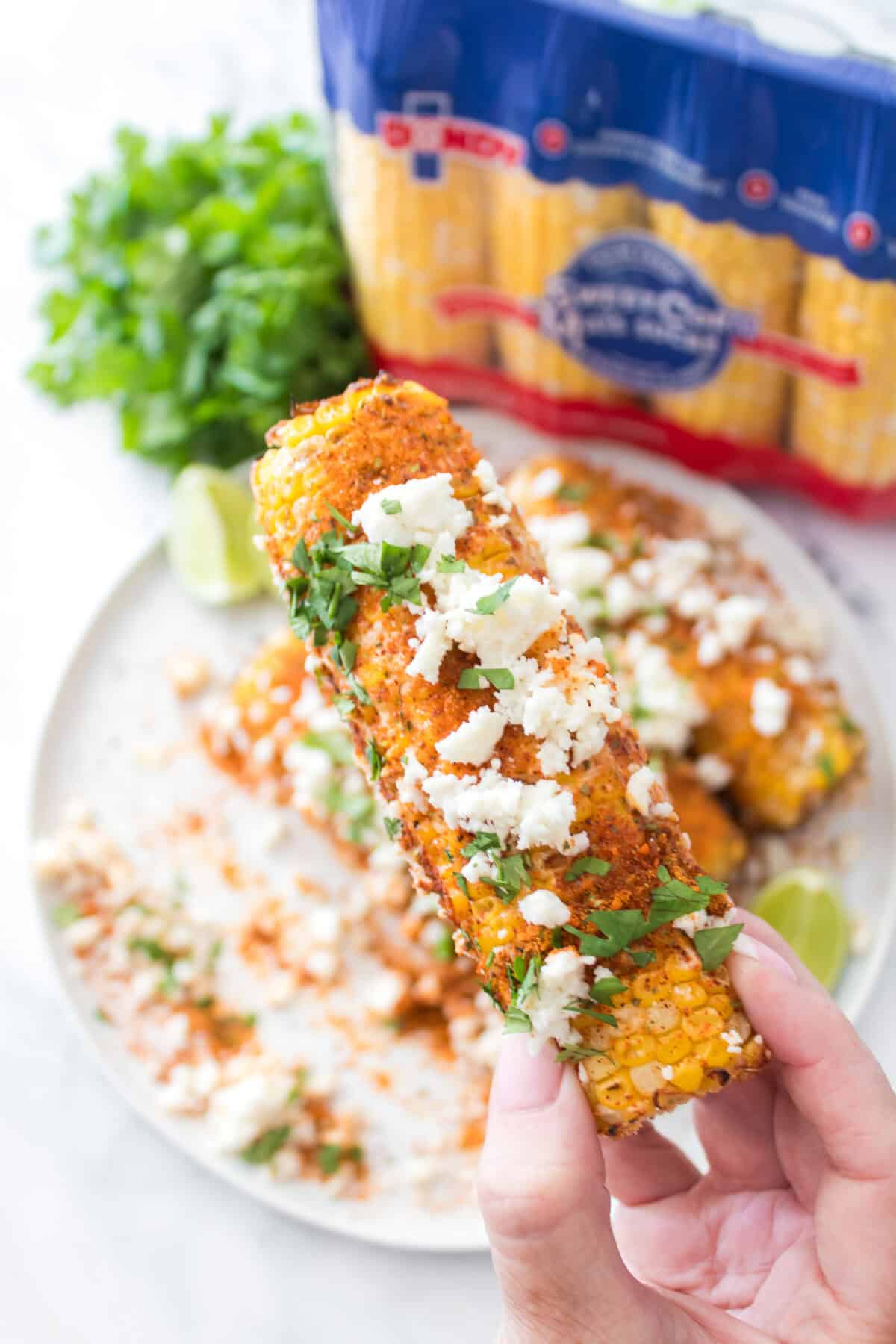 hand holding elote corn over the top of a plate of elote corn