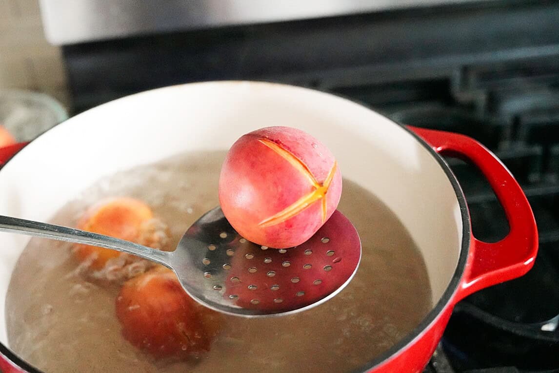 Peaches in boiling water with a slotted spoon holding one peach