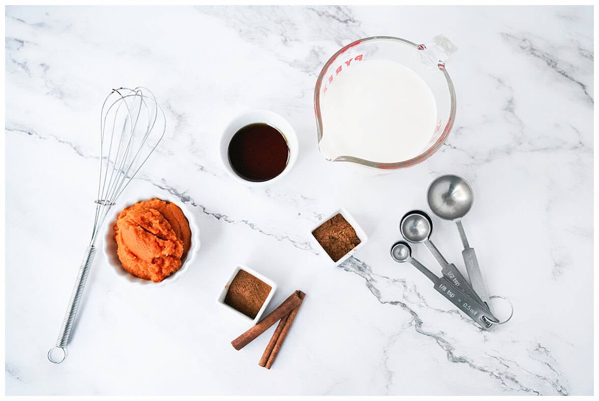 overhead shot of ingredients to make pumpkin spice coffee creamer: Whole milk in measuring glass, whisk, small white bowl with pumpkin puree, cinnamon, nutmeg, cinnamon sticks and syrup
