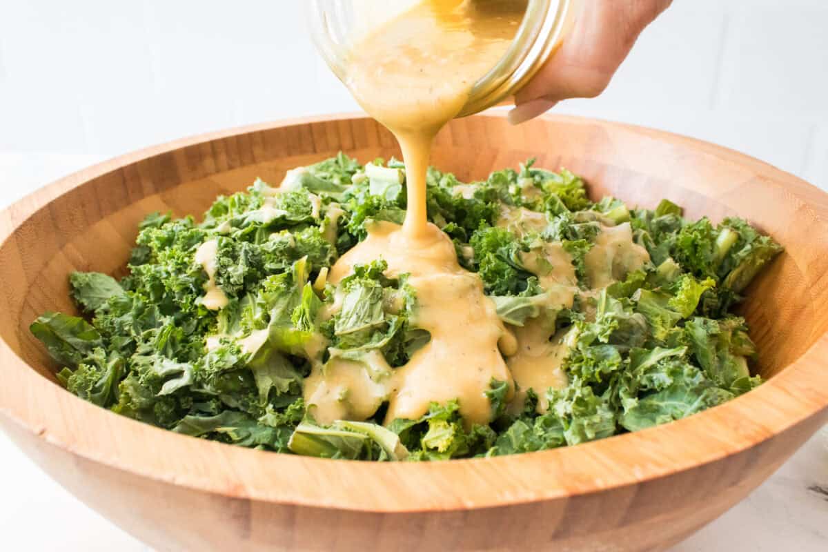 hand pouring tahini dressing over freshly chopped kale in bamboo salad bowl