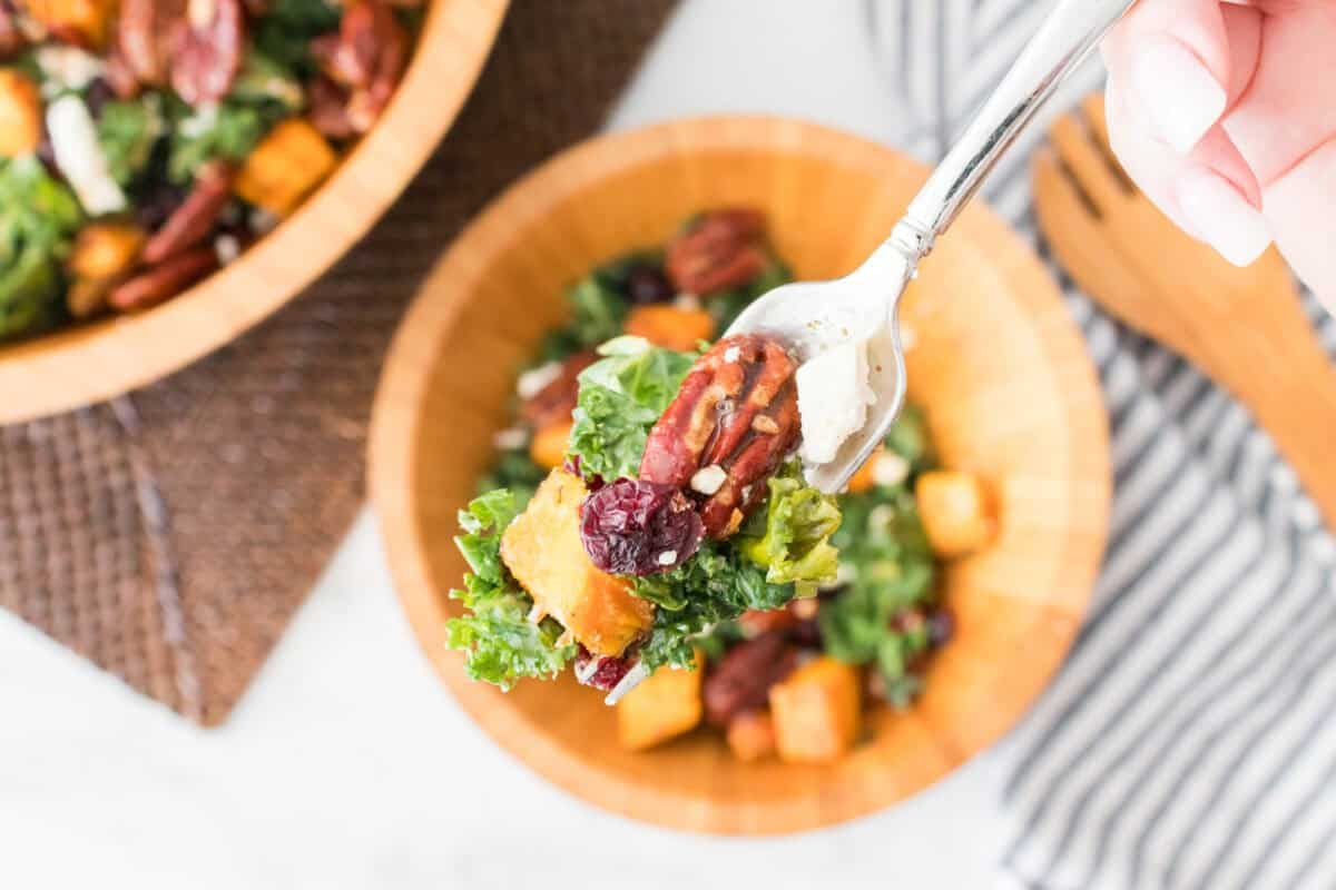 hand holding fork with a bite of sweet potato kale salad with candied pecans, feta cheese and dried cranberry