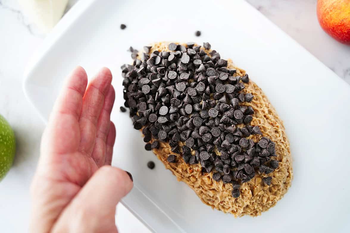hand placing mini chocolate chip morsels over the top of apple toffee dip shaped into the form of a football on white serving platter