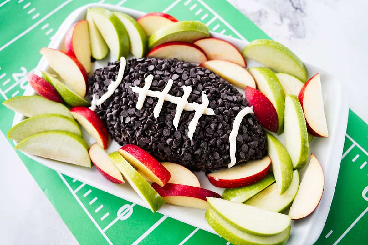 45 degree shot of apple toffee dip on white serving platter with dip shaped as a football, coated in chocolate chips and white icing for laces on the football, served with slices of apples