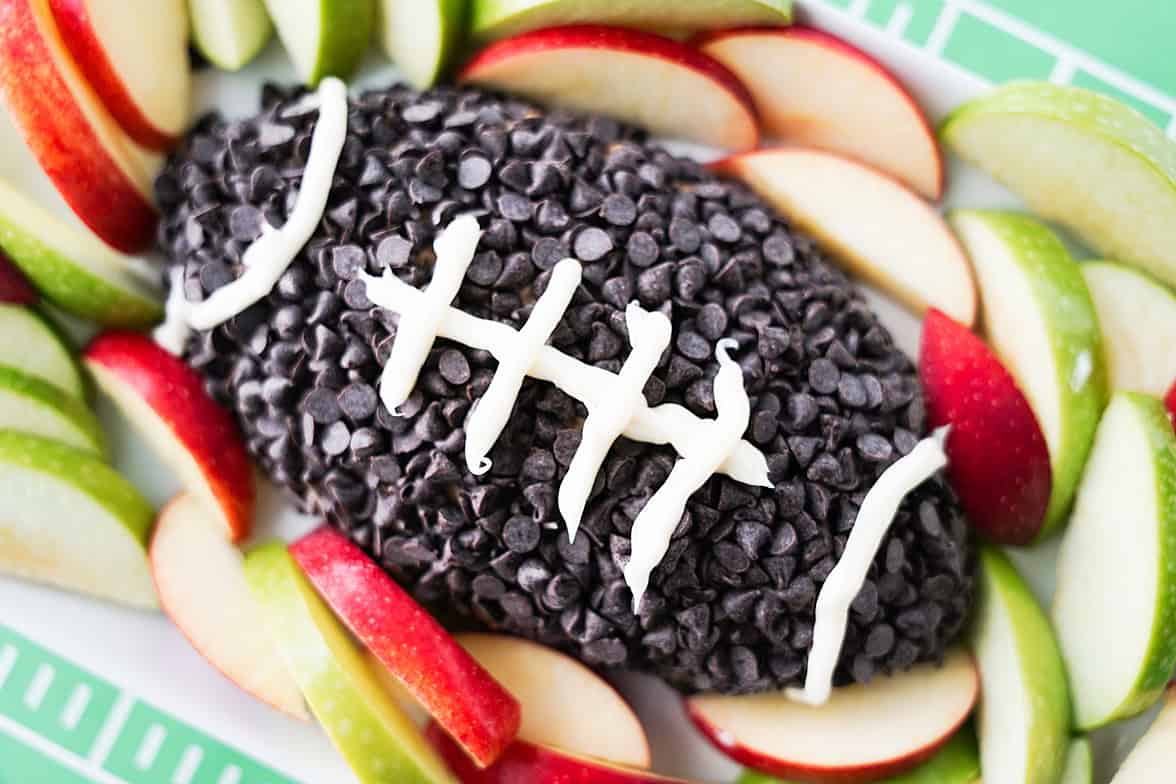 apple toffee dip shaped into a football, coated in chocolate chips and white icing for lines in the football on serving tray surrounded with apple slices