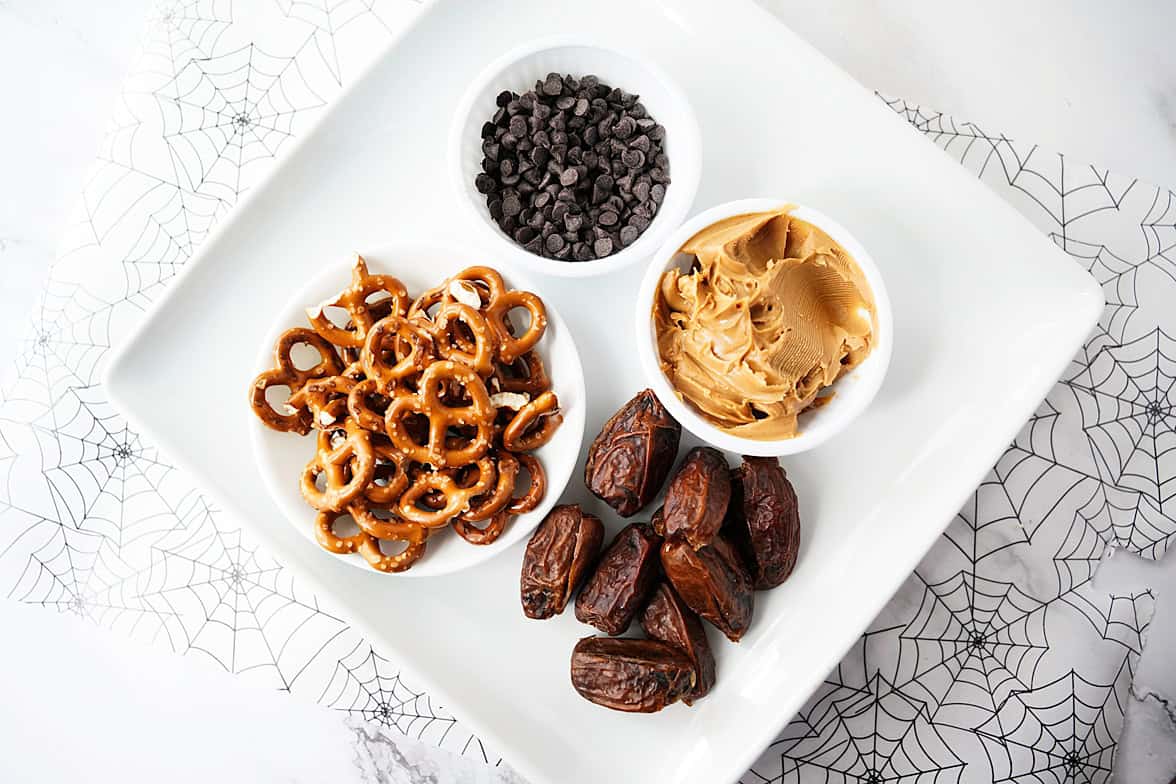overhead shot of ingredients to make date spiders: plate of pretzel twists, bowl of mini chocolate chips, bowl of peanut butter and dates on white serving platter