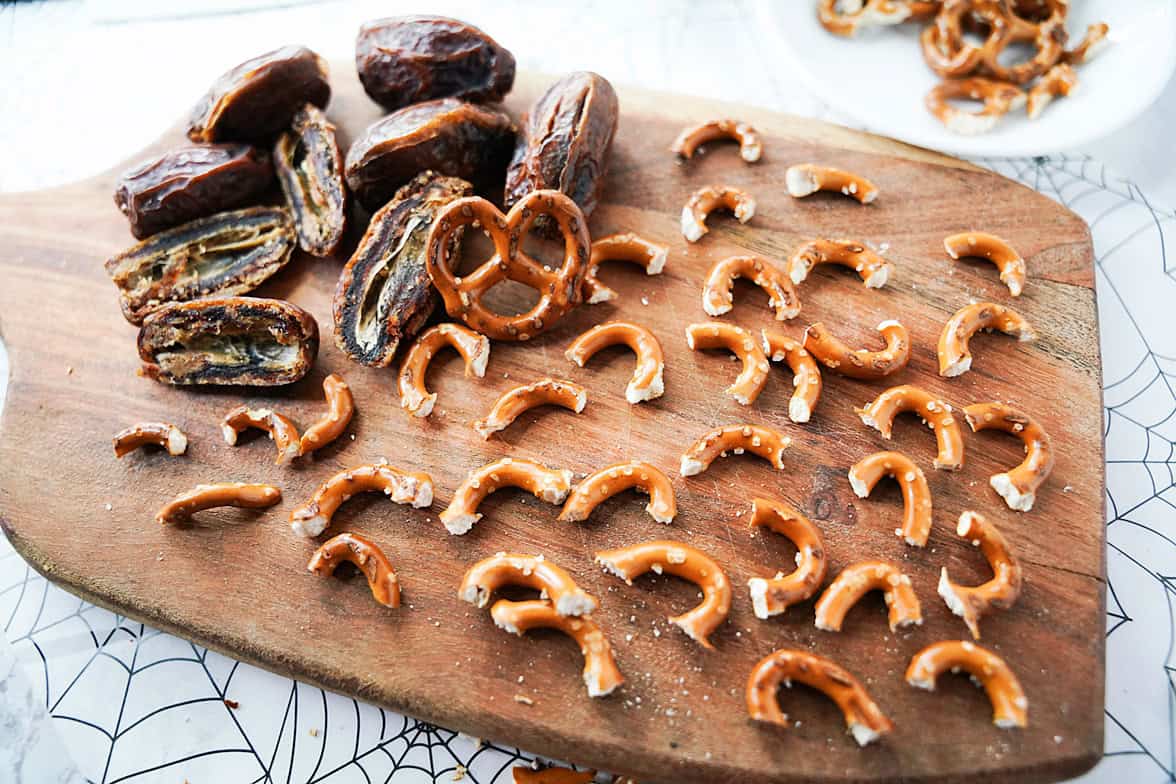 dates cut in half on cutting board with pretzel twists broken into pieces to create legs for a spider