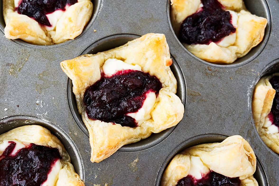close up shot of muffin tin with puff pastry that has been brushed with egg wash, with cream cheese mixture and plum jelly in each puff pastry after baking