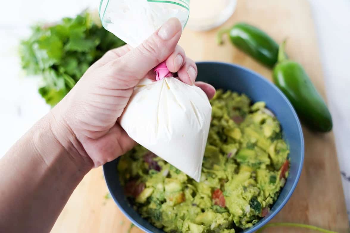 Bowl of guacamole with a plastic bag of sour cream overhead for piping the web