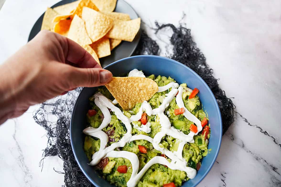 overhead shot of spiderweb gauacamole with hand dipping a chip from bowl in top left corner