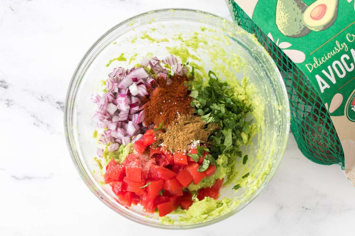 overhead shot of all ingredients to make homemade guacamole in glass bowl before mixing: cilantro, tomato, 1/2 red onion, bag of westfalia fruit avocados, salt, chili powder, cumin, garlic salt and lime juice