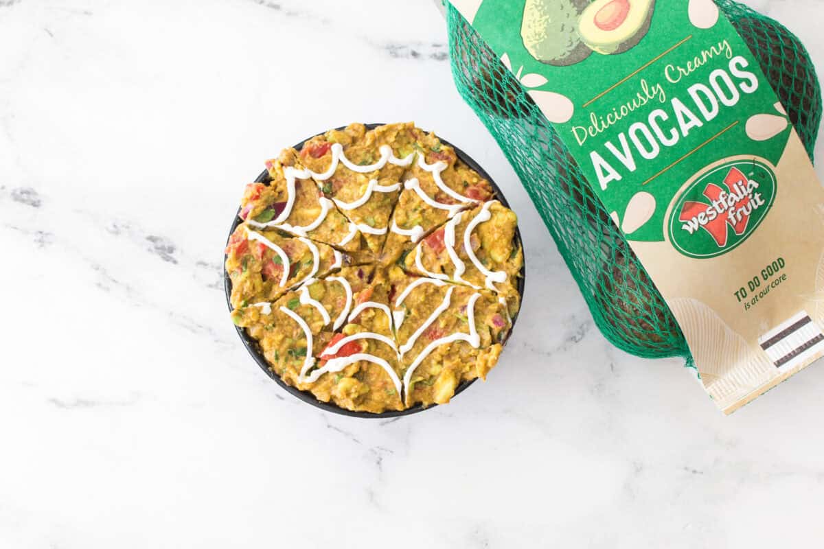 overhead shot of guacamole with sour cream on top design to look like a spiderweb with bag of westfalia fruit avocados