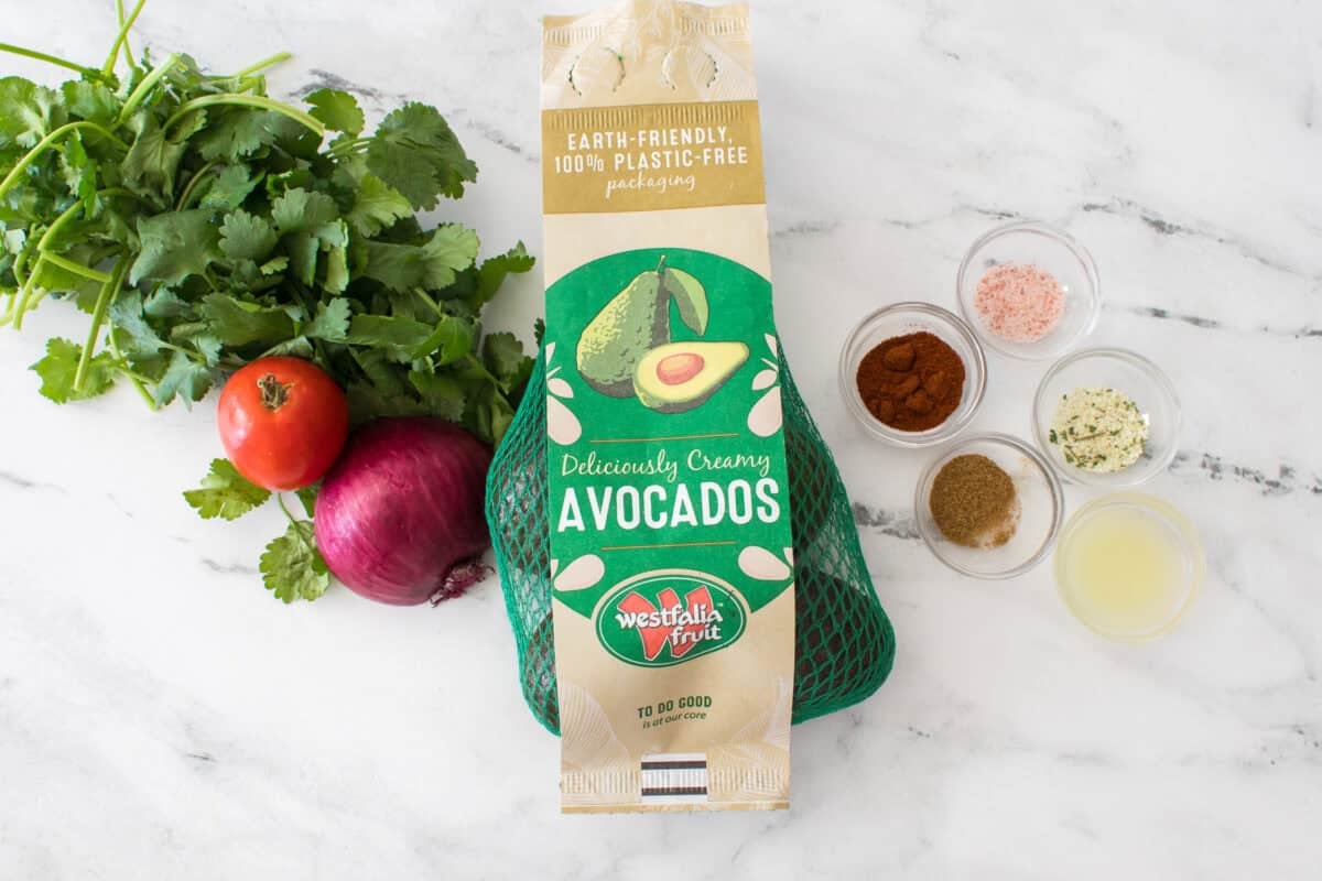 overhead shot of ingredients to make homemade guacamole: cilantro, tomato, 1/2 red onion, bag of westfalia fruit avocados, salt, chili powder, cumin, garlic salt and lime juice