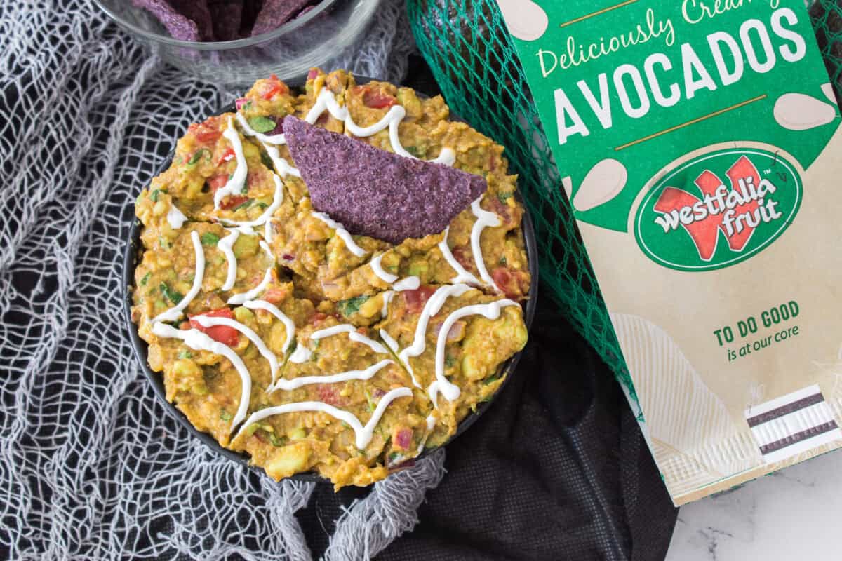 overhead shot of spiderweb guacamole with bag of westfalia fruit avocados with a black tortilla chip in guacamole