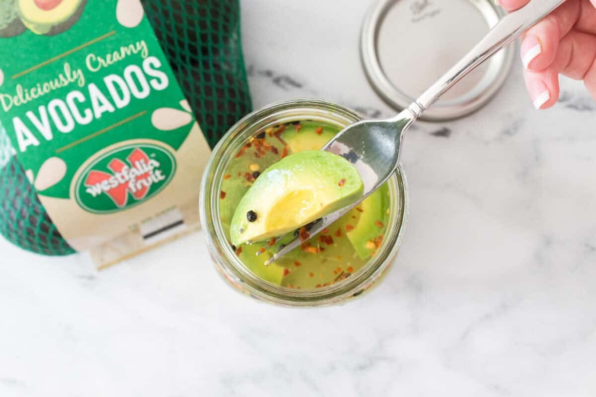 bag of westfalia fruit avocados with avocado cut in half and big mouth mason jar with pickled avocados and fork grabbing a pickled avocado out