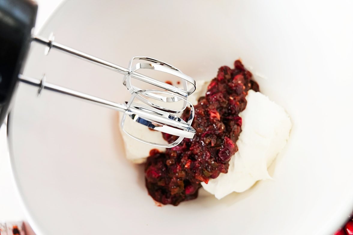 white bowl with block of cream cheese and cranberry jalapeno jam mixture with hand mixture over the bowl before combining