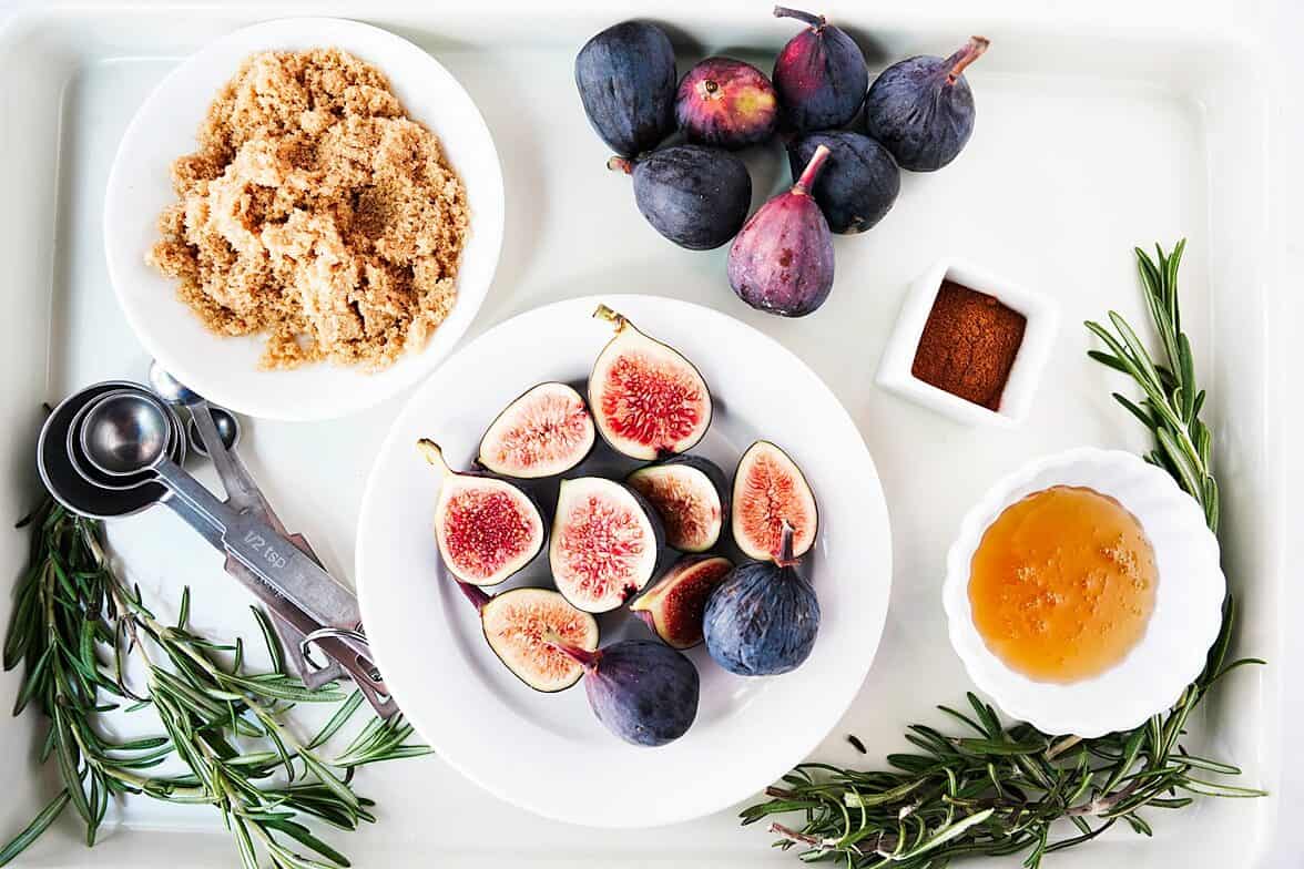 Ingredients for oven roasted figs on a white tray: figs, brown sugar, cinnamon, honey and rosemary sprigs