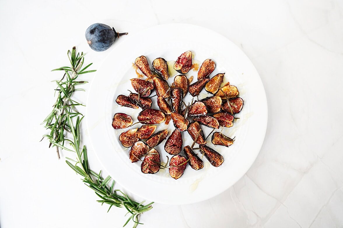 Overhead shot of oven roasted figs on a white play surrounded by rosemary and a fresh fig