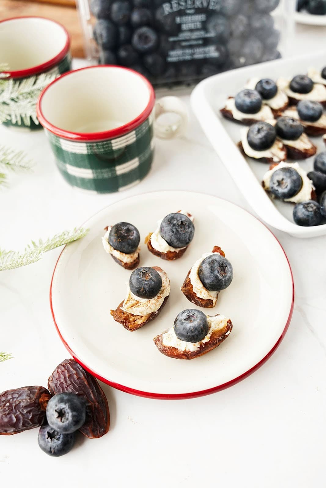 Vertical images of blueberry mascarpone stuffed dates ona. white plate with green + red coffee cup