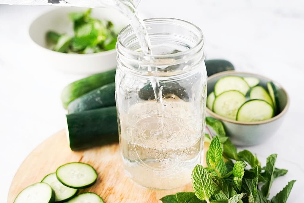 pouring water into a mason jar surrounded by cucmbers and mint