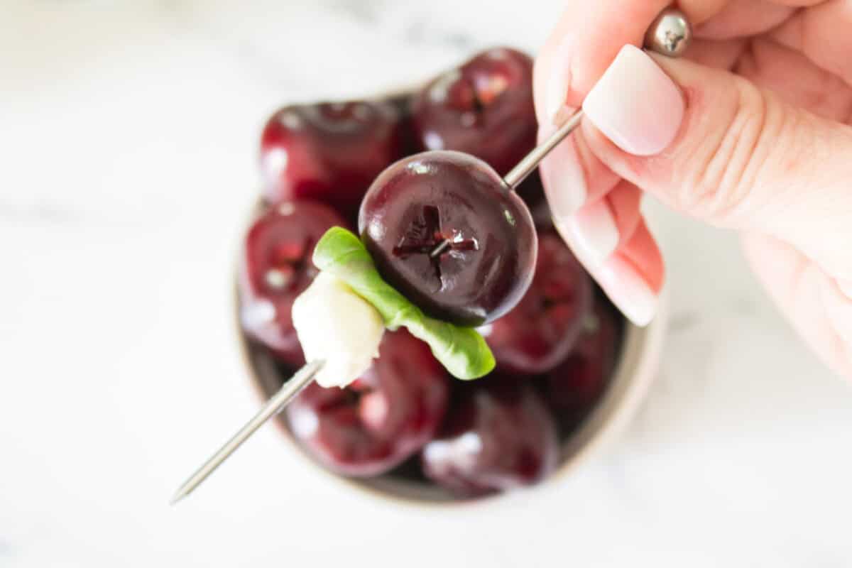 A close-up, top-down shot of a hand holding a metal cocktail skewer threaded with a whole pitted dark cherry, a folded fresh green basil leaf, and a small white mozzarella pearl over a bowl of fresh cherries.