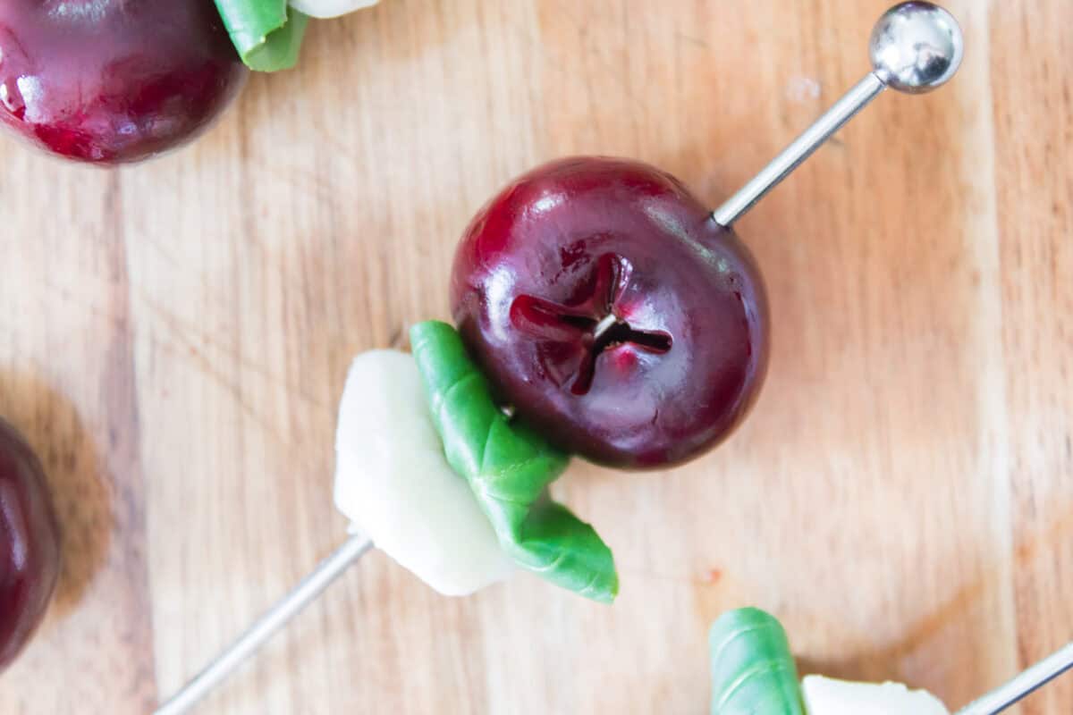 A close-up, slightly high-angle shot focusing on the assembly of a sweet cherry caprese skewer, showing a metal cocktail pick halfway through piercing a whole pitted cherry, a fresh green basil leaf, and a white mozzarella pearl on a wooden cutting board.