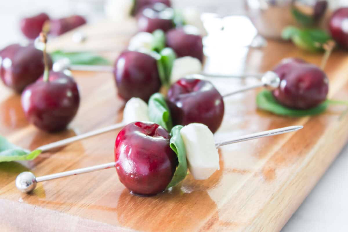 A slightly elevated, side-angle shot of several sweet cherry caprese skewers lined up across a wooden serving board, showcasing a clear view of the entire spread of red cherries, folded basil leaves, and white mozzarella pearls before the balsamic glaze is drizzled.