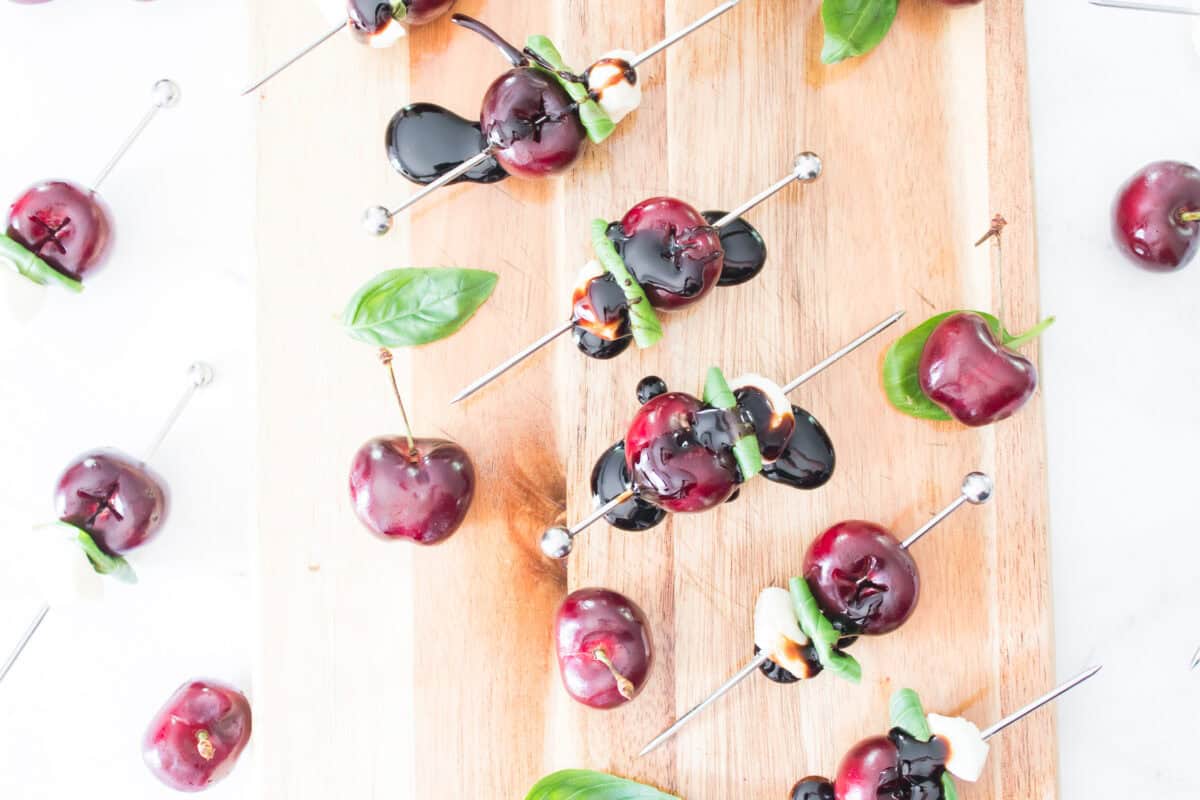 A top-down flat lay shot of sweet cherry caprese skewers scattered across a wooden serving board on a white marble surface, showing the skewers heavily drizzled with rich, dark balsamic glaze alongside loose whole cherries and fresh basil leaves.