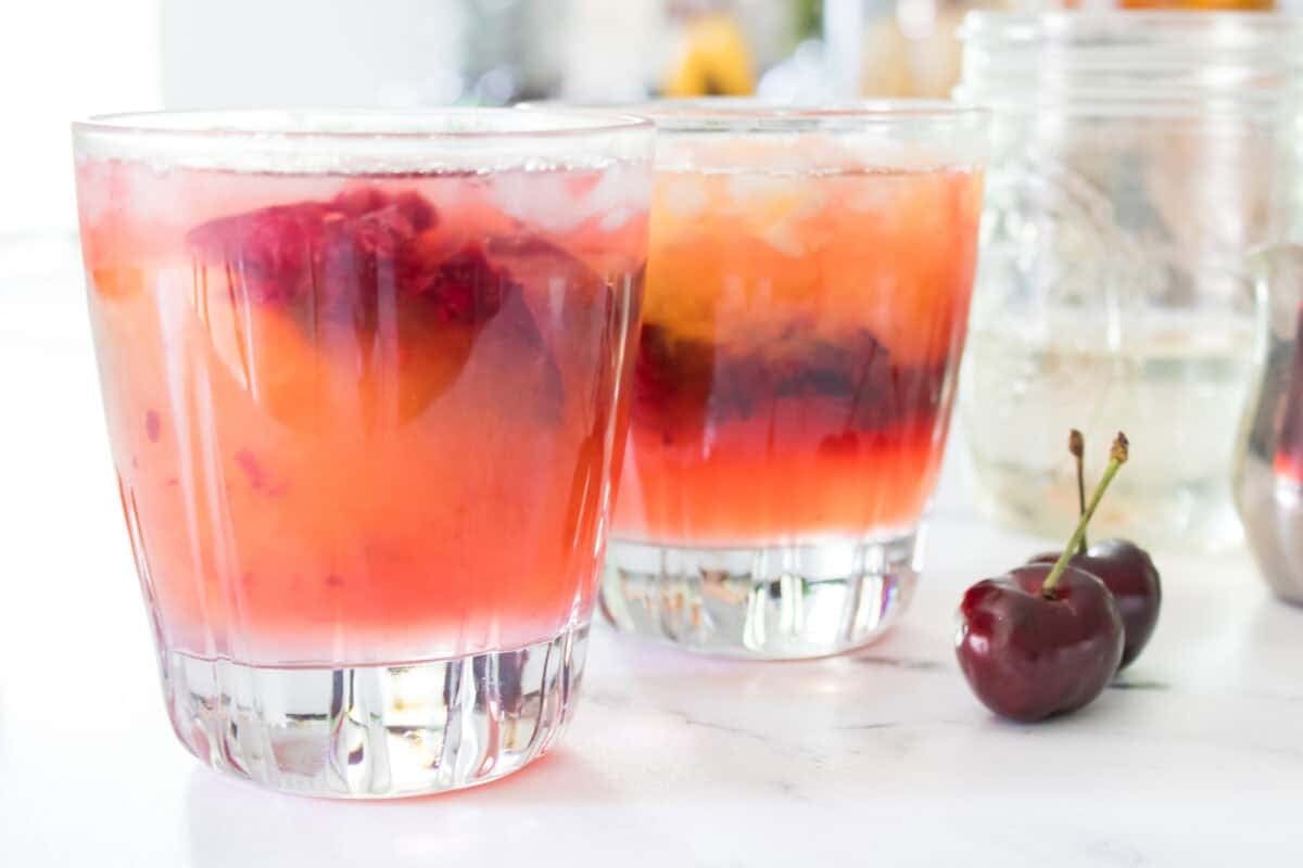 A side-angle shot of two cherry mango margaritas in lowball glasses filled with crushed ice, displaying a beautiful ombre effect with deep red cherry juices settled at the bottom and a vibrant peach-colored mango layer on top, with fresh whole cherries resting on the counter in front.