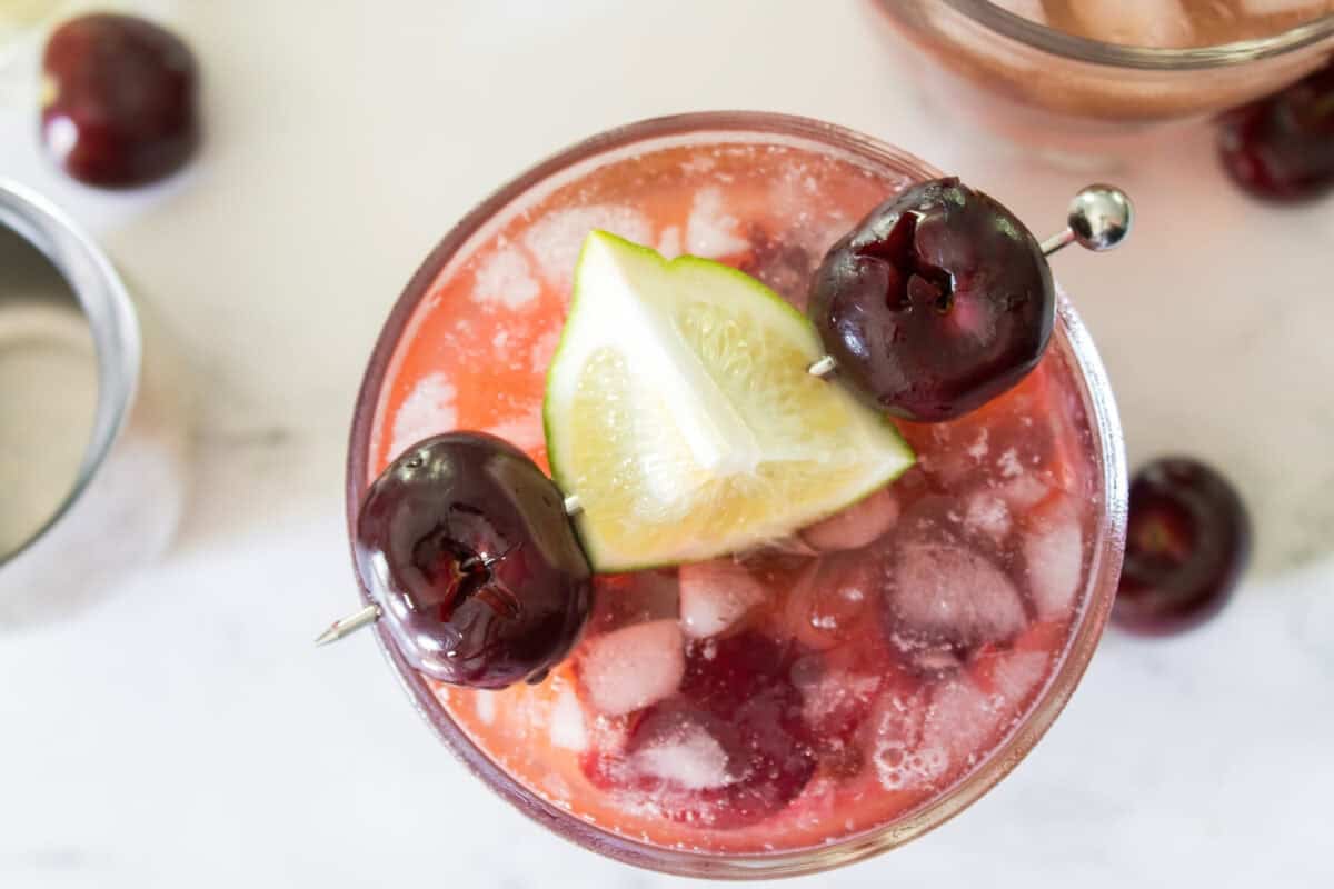 A close-up, top-down shot looking directly into a clear glass filled with a cherry mango margarita and crushed ice, topped with a metal cocktail skewer holding two whole, dark sweet cherries and a fresh green lime wedge.