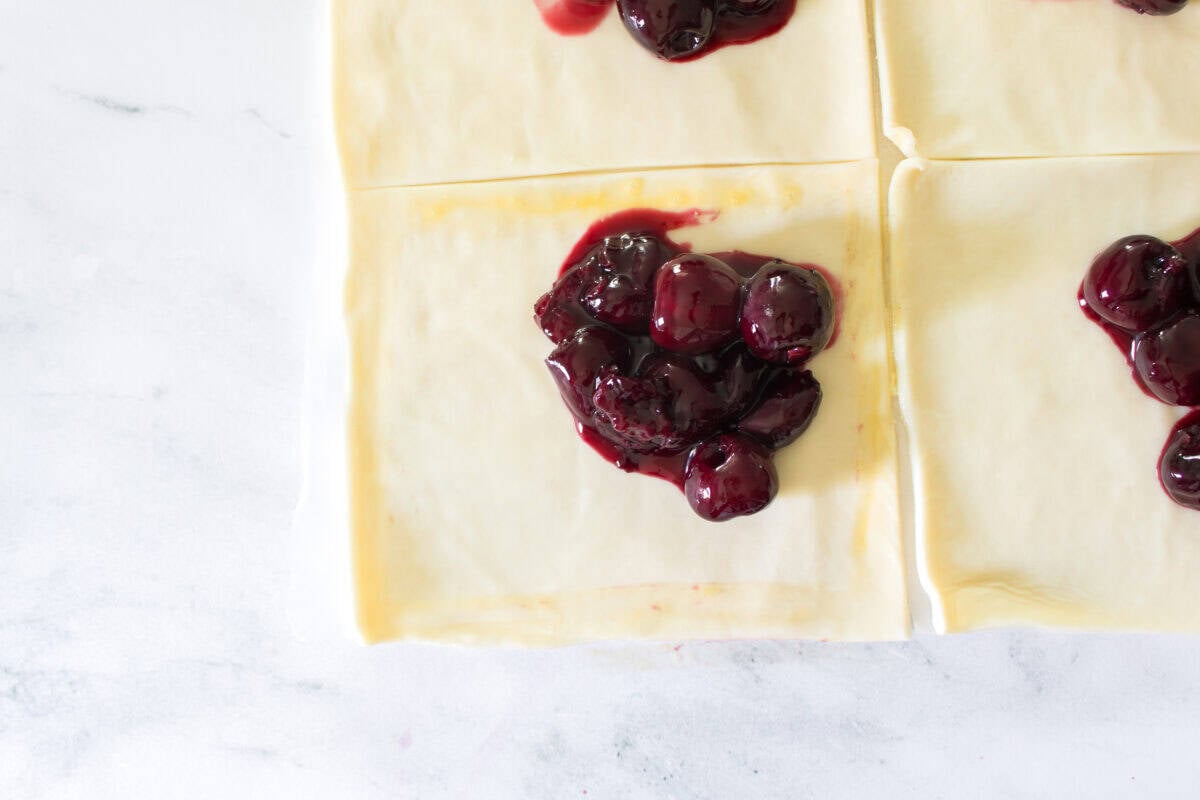 An overhead shot showing a square of uncooked puff pastry dough on a marble surface with a spoonful of glossy, dark cherry filling placed neatly in the center, ready to be folded into a turnover.